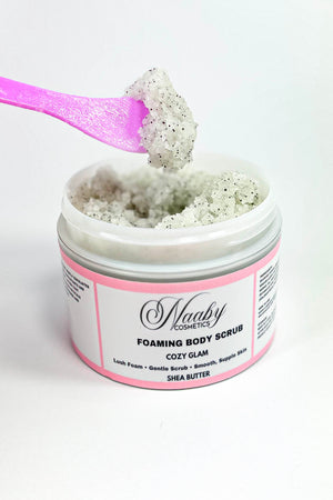 Foaming body scrub in a jar with a pink spoon lifting some scrub above it on a white background.