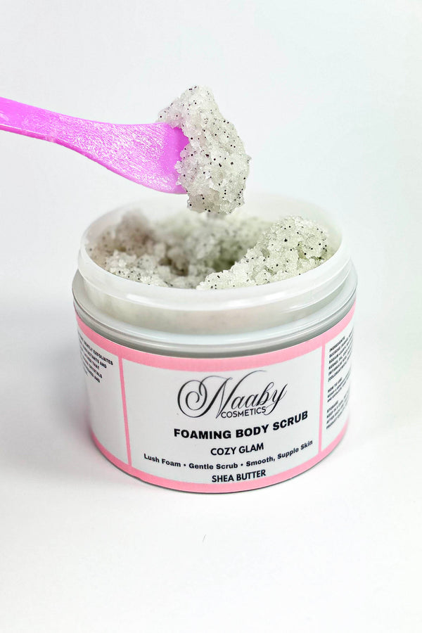 Foaming body scrub in a jar with a pink spoon lifting some scrub above it on a white background.
