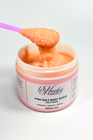 Pink salt body scrub being scooped out of a container by a purple spoon on a white background.Cozy Glam – Pink Salt Scrub