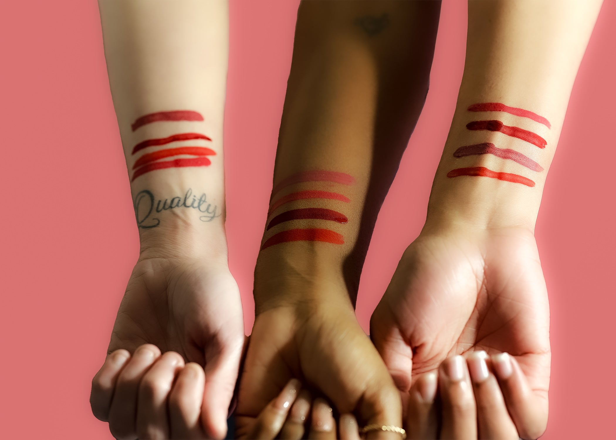 Three hands with red makeup swatches on wrists against a pink background